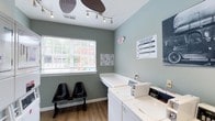 This laundry facility offers a clean, well-maintained environment designed with both functionality and comfort in mind. The walls are painted in a calming sage green tone, creating a pleasant atmosphere for residents during their visits. Natural light filters into the space through a window fitted with horizontal blinds, brightening the room and adding an open, airy feel.

The facility is equipped with top-load washing machines from the Quantum series, with posted instructions and washer options clearly displayed on the wall for user reference. Adjacent signage provides additional service information, ensuring residents have easy access to operational guidance. A white folding or utility counter sits atop the machines, offering a convenient surface for sorting or folding laundry.

Track lighting mounted on the ceiling provides ample illumination throughout the space, complemented by a decorative ceiling accent. Wood-look flooring runs throughout, adding a warm and polished finish to the utilitarian setting. Two black chairs are positioned along the wall beneath the window, offering a comfortable waiting area. The overall layout is compact yet efficiently organized, making the most of the available space.