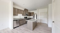 This single-floor unit presents a cohesive, modern design throughout its living spaces. The kitchen serves as a central highlight, featuring rich medium-toned cabinetry with brushed nickel hardware, granite countertops, and a full suite of stainless steel appliances including a refrigerator, electric range, over-the-range microwave, and dishwasher. A center island with matching granite provides additional workspace and casual dining potential. Light-toned luxury vinyl plank flooring runs throughout the kitchen and main living areas, contributing to a clean, contemporary aesthetic.

The bedroom offers a comfortable, carpeted environment with neutral walls and a window framing an outdoor view of trees and what appears to be a pond, providing a pleasant natural backdrop. Storage is well-addressed through a generously sized walk-in closet equipped with white wire shelving systems and carpeted flooring.

The bathroom maintains design continuity with the kitchen, featuring the same medium-toned cabinetry, granite countertop, an undermount sink with brushed nickel fixtures, a large mirror, and neutral tile flooring. The overall palette of the unit—whites, grays, and warm browns—creates a consistent and inviting atmosphere throughout.