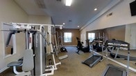 This fitness center is thoughtfully designed to accommodate a wide range of exercise needs within a single, open floor layout. The space is outfitted with an impressive selection of equipment, including treadmills, elliptical machines, stationary bikes, and a multi-station strength training unit, providing options for both cardiovascular and resistance training. The room features a neutral color palette with warm beige walls and carpeted flooring, contributing to a comfortable and focused workout atmosphere. Recessed lighting and overhead fluorescent fixtures ensure the space is well-lit throughout. Large windows along one wall invite natural light and offer a view of the outdoors, enhancing the overall ambiance. Full-length mirrors are mounted along an adjacent wall, a practical addition that allows users to monitor their form and visually expand the sense of space. Ventilation is addressed with ceiling vents positioned throughout the room. The layout is open and uncluttered, allowing for easy movement between equipment. A glass-paned door provides a secondary access point, adding to the room's accessibility and airflow. Overall, this fitness center is a well-organized, fully functional space suited for a consistent and varied exercise routine.