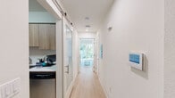 This efficiently designed single-floor unit offers a complete and functional living experience within a compact footprint. Light wood flooring runs throughout, creating a warm, cohesive aesthetic that ties each area together seamlessly.

A bright, central hallway connects the main spaces and features white walls, recessed lighting, and a smart home control panel, reflecting the unit's modern sensibility. A sliding barn-style glass door provides stylish access to the kitchen, which is outfitted with floor-to-ceiling wood-tone cabinetry, white quartz countertops, and a full suite of stainless steel appliances including a gas range, over-the-range microwave, and dishwasher — a fully equipped space well-suited for everyday cooking.

The living area is clean and minimalist, with white walls and ample room for a full-sized sofa, offering a calm and uncluttered retreat. Adjacent to this is a sleeping area that comfortably accommodates a double bed alongside a closet with louvered doors, providing practical storage.

The bathroom is finished with a wood-grain vanity, backlit mirror, white quartz countertop, and a glass-enclosed shower-tub combination, delivering a polished, contemporary feel. Natural light filters in from the far end of the hallway, lending the space an open and airy quality throughout.
