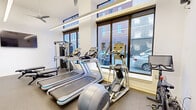 Located at 142 W York St in Norfolk, VA, this contemporary fitness center offers a well-appointed workout environment designed to support a full range of exercise routines. The space is housed on a single floor and makes efficient use of its compact footprint, delivering a comprehensive gym experience without sacrificing functionality or comfort.

Large floor-to-ceiling windows line one side of the room, flooding the interior with abundant natural light and offering views of the surrounding urban streetscape. Linear LED lighting runs along the ceiling, complementing the bright, white walls and creating a clean, energizing atmosphere. A ceiling fan adds to the air circulation throughout the space.

The fitness floor is outfitted with durable rubber flooring suitable for high-impact activity. Cardio equipment includes multiple treadmills, an elliptical machine, and a Precor spin bike. A multi-station cable machine provides versatile strength training options, while a well-stocked dumbbell rack, medicine balls, stability balls, and a plyometric box support functional and free-weight training. A flat-screen TV is mounted on the wall for entertainment during workouts.

A glass-paned door with black steel framing connects the fitness room to an adjacent lounge area, adding an architectural touch while maintaining a sense of openness. The overall design balances modern aesthetics with practical utility, making this fitness center a standout amenity.