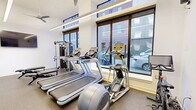 Located at 142 W York St in Norfolk, VA, this contemporary fitness center offers a well-appointed workout environment designed to support a full range of exercise routines. The space is housed on a single floor and makes efficient use of its compact footprint, delivering a comprehensive gym experience without sacrificing functionality or comfort.

Large floor-to-ceiling windows line one side of the room, flooding the interior with abundant natural light and offering views of the surrounding urban streetscape. Linear LED lighting runs along the ceiling, complementing the bright, white walls and creating a clean, energizing atmosphere. A ceiling fan adds to the air circulation throughout the space.

The fitness floor is outfitted with durable rubber flooring suitable for high-impact activity. Cardio equipment includes multiple treadmills, an elliptical machine, and a Precor spin bike. A multi-station cable machine provides versatile strength training options, while a well-stocked dumbbell rack, medicine balls, stability balls, and a plyometric box support functional and free-weight training. A flat-screen TV is mounted on the wall for entertainment during workouts.

A glass-paned door with black steel framing connects the fitness room to an adjacent lounge area, adding an architectural touch while maintaining a sense of openness. The overall design balances modern aesthetics with practical utility, making this fitness center a standout amenity.