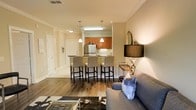 This single-floor space showcases an open-concept layout that connects the living area and kitchen in a cohesive, flowing design. The living area features hardwood-style flooring, neutral beige walls accented with crown molding, and is furnished with a contemporary gray sofa accessorized with blue accent pillows. A stylish floor lamp with a dark shade and decorative sculptural elements adds a refined touch to the space.

The kitchen is positioned at the rear and is highlighted by warm medium-tone wood cabinetry, a tasteful tile backsplash, and stainless steel appliances, including a refrigerator. Pendant lights hang above a dark-topped kitchen island or bar counter, which is lined with upholstered bar-height chairs, creating an inviting spot for casual dining or entertaining.

A hallway extends from the main living area, leading to additional rooms and featuring a white paneled door that contributes to the clean, classic aesthetic. The warm, ambient lighting throughout the space enhances its welcoming and well-appointed atmosphere, making it both functional and visually appealing.