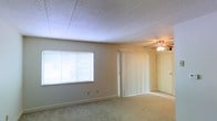 This single-floor space features a main living area finished with wall-to-wall carpet that adds a soft, comfortable feel underfoot. The room is illuminated by natural light streaming through a horizontal window fitted with white blinds, allowing for adjustable light control throughout the day. A ceiling fan with an integrated light fixture is centrally positioned, providing both air circulation and ambient lighting. The textured tile ceiling adds a classic architectural detail overhead. Vertical blinds cover what appears to be a sliding door, offering access to an adjacent area or exterior while maintaining privacy. The neutral-toned walls and clean baseboards give the space a simple, functional aesthetic. The overall layout is open and unobstructed, providing flexibility in how the room can be arranged and utilized.