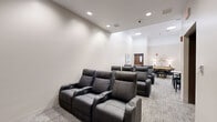 Located at 797 Main St in Hartford, Connecticut, this single-floor lounge and entertainment room offers a thoughtfully designed space for relaxation and social interaction. The room is finished with neutral, light-toned walls and textured low-pile carpeting that lends a contemporary yet comfortable atmosphere throughout.

The seating area anchors one side of the room with rows of premium dark leather recliner chairs arranged in theater-style fashion, each equipped with built-in cup holders and power reclining functionality. This configuration makes the area well-suited for screenings or communal viewing. A large-screen display and wood-paneled accent wall are visible at the front of the seating arrangement, further reinforcing the cinematic feel.

Toward the opposite end of the room, a dedicated gaming area features a round poker table with inlaid felt surface, surrounded by sleek modern dining chairs. A small bistro table and additional seating provide an intimate nook nearby. The walls in this zone are adorned with a set of four textured white canvas panels, adding an artistic, gallery-like element to the space.

Recessed ceiling lighting is installed throughout, providing even, well-distributed illumination. A bold black abstract painting on one wall adds a striking visual accent. The overall design balances functionality and style, creating an inviting multi-use social environment.