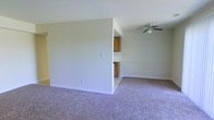 This single-floor residence at Brookwood Apartments offers a straightforward, functional layout designed for comfortable everyday living. The main living area is spacious and open, featuring wall-to-wall carpeting in a neutral tone that adds warmth underfoot. Light, neutral-painted walls keep the space feeling bright and versatile, ready to accommodate a variety of interior styles. A ceiling fan is installed overhead, providing both airflow and a subtle decorative element to the room. A partial dividing wall separates the living area from the kitchen, creating a semi-open concept that maintains a sense of connection between the two spaces while offering a degree of visual separation. The kitchen features warm wood cabinetry, offering ample storage for everyday needs. Clean white trim and baseboards run throughout, adding a crisp, finished look to the interior. The overall ambiance is clean, neutral, and move-in ready, making it well-suited for those looking for a simple, low-maintenance living space within the Brookwood Apartments community.