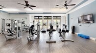 Located at 940 W Canton Ave in Winter Park, Florida, this fitness center offers a generously sized, single-floor workout facility designed to support a comprehensive range of exercise needs. The space features rich hardwood-style flooring throughout, paired with crisp white walls accented by a distinctive blue trim that runs along the upper perimeter, lending the interior a clean and modern aesthetic.

The facility is well-organized into distinct training zones. The strength training area is outfitted with a variety of Life Fitness selectorized machines, a Smith machine with a barbell bench setup, and adjustable weight benches, all positioned alongside floor-to-ceiling mirrors that enhance the sense of space. A dedicated free weights and functional training section includes a medicine ball rack, stability balls, a step platform, and a water cooler for convenience.

The cardio zone is positioned toward the front of the room, bathed in natural light streaming through a row of large glass French doors that offer pleasant outdoor views, creating an inviting and energizing atmosphere. Treadmills, ellipticals, and a recumbent bike are among the cardio options available.

Overhead, a drop tile ceiling with recessed fluorescent lighting and multiple ceiling fans ensures even illumination and comfortable air circulation throughout. Wall-mounted flat-screen televisions are positioned at key locations for entertainment during workouts, completing this thoughtfully designed fitness environment.