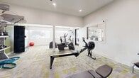Located at 8250 E Golf Links Rd in Tucson, Arizona, this fitness center offers a thoughtfully designed exercise space contained within a single open room. The interior features a clean, modern aesthetic with white walls accented by a bold olive-green feature wall, and a patterned carpet in neutral gray and yellow tones that adds visual energy underfoot. Recessed lighting and a ceiling fan with sleek, dark blades provide both illumination and air circulation throughout the space.

The room is comprehensively outfitted for a full-body workout experience. Cardio equipment includes a treadmill, elliptical trainers, a stationary cycle, and a Concept2 RowERG rowing machine. For strength training, the facility provides a multi-function cable machine, a rack of dumbbells spanning multiple weight increments paired with an adjustable bench, a set of kettlebells, and battle ropes for functional fitness. A wall-mounted flat-screen television is positioned in the cardio area for entertainment during workouts.

Natural light filters in through several windows fitted with blinds, keeping the space bright and open. A glass-panel door connects to an adjacent area, enhancing the sense of flow. The layout efficiently maximizes the available floor area, ensuring a variety of training options without feeling cramped, making this a versatile and well-appointed fitness facility.