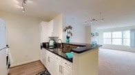 This single-floor residence offers a thoughtfully designed open-concept layout that connects the kitchen and living area in a cohesive, functional arrangement. The kitchen is a standout feature, showcasing crisp white cabinetry with sleek silver hardware, rich dark granite countertops, and a peninsula that doubles as a breakfast bar — perfect for casual dining or entertaining. A stainless steel sink with a modern faucet is integrated into the peninsula, adding both style and practicality. Track lighting overhead illuminates the space with a contemporary touch, while light wood-style flooring grounds the kitchen area with warmth and durability. The open pass-through design creates a natural sightline into the adjacent living area, which features plush carpeting and bright, natural light streaming in through a large window. The overall ambiance is clean and inviting, with a neutral color palette of white walls and soft tones that provide a versatile backdrop. The seamless transition between the kitchen and living space makes this layout ideal for everyday living, offering both comfort and a modern aesthetic throughout.