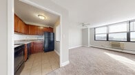 This single-floor residential unit offers a practical and comfortable layout designed for everyday living. The kitchen is equipped with warm-toned wood cabinetry, granite-style countertops, a black refrigerator, a stainless steel dishwasher, and a black range, all set on durable tile flooring. A pass-through window connects the kitchen to the main living area, enhancing the open feel between the two spaces. The living room is generously sized, featuring plush wall-to-wall carpeting, a ceiling fan, a baseboard heating system, and a large multi-pane window that fills the room with natural light and offers views of the surrounding cityscape. A wall-mounted air conditioning unit ensures year-round comfort. The bedroom continues the carpeted aesthetic and includes a wide set of louvered bi-fold closet doors providing substantial storage space. It also features its own window with an outdoor view and a baseboard heating unit. Throughout the unit, neutral gray-toned walls and clean white trim create a bright, cohesive atmosphere. The entry door leads directly into the open living space, making for a seamless and efficient floor plan.