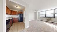This single-floor residential unit offers a practical and comfortable layout designed for everyday living. The kitchen is equipped with warm-toned wood cabinetry, granite-style countertops, a black refrigerator, a stainless steel dishwasher, and a black range, all set on durable tile flooring. A pass-through window connects the kitchen to the main living area, enhancing the open feel between the two spaces. The living room is generously sized, featuring plush wall-to-wall carpeting, a ceiling fan, a baseboard heating system, and a large multi-pane window that fills the room with natural light and offers views of the surrounding cityscape. A wall-mounted air conditioning unit ensures year-round comfort. The bedroom continues the carpeted aesthetic and includes a wide set of louvered bi-fold closet doors providing substantial storage space. It also features its own window with an outdoor view and a baseboard heating unit. Throughout the unit, neutral gray-toned walls and clean white trim create a bright, cohesive atmosphere. The entry door leads directly into the open living space, making for a seamless and efficient floor plan.