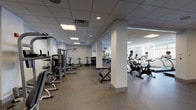 This comprehensive fitness center is thoughtfully organized across a single floor, offering a wide variety of workout zones to accommodate diverse training needs. The space features a clean, modern aesthetic with neutral grey walls, a suspended grid ceiling with recessed and panel lighting, and durable speckled rubber flooring throughout — designed for both performance and easy maintenance.

The cardio zone is generously appointed with a row of treadmills, stationary upright and recumbent bikes, and elliptical machines, positioned near windows that allow natural light to filter into the space. Full-length mirrors line the walls, enhancing the sense of openness. A dedicated strength training area houses a series of cable and resistance machines arranged in a structured layout, supporting targeted muscle group training. A free weight section features a dumbbell rack alongside adjustable benches, catering to both beginners and experienced lifters.

A separate functional training zone provides open floor space equipped with medicine ball racks, stability balls, and foam rollers, ideal for warm-ups, stretching, or dynamic workouts. Structural columns throughout the space define natural zones without obstructing flow. An additional room, visible through a glass-panel door, appears to house supplementary cardio equipment. The overall ambiance is professional, clean, and well-maintained, making it a fully capable facility for a complete fitness routine.