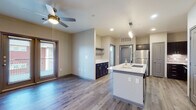 This thoughtfully designed single-floor residence offers a seamless blend of contemporary style and everyday functionality. The open-concept layout connects the kitchen and living area, creating a bright, cohesive space anchored by durable wood-look flooring throughout. A ceiling fan with a brass-accented light fixture adds both comfort and a touch of warmth to the living area, which benefits from natural light streaming in through a large window with blinds.

The kitchen is a standout feature, showcasing a central island with a white quartz countertop and an undermount sink fitted with a sleek pull-down faucet. Dark espresso cabinetry provides rich contrast against the light countertops, while a classic subway tile backsplash adds texture. Stainless steel appliances, including a refrigerator, range, and over-the-range microwave, complete the well-equipped cooking space. A pendant light cluster above the island adds an elegant finishing touch. A set of double doors suggests generous pantry or closet storage nearby.

The bedroom is a calm retreat, featuring plush wall-to-wall carpeting, a ceiling fan, and multiple windows — including elevated transom-style windows — that invite abundant natural light.

The bathroom is finished with light wood-look flooring, a wide vanity with dark cabinetry, a crisp white countertop, a large mirror, and a full tub and shower accented by large-format neutral tile surround.