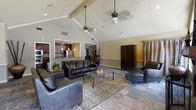 This single-story space is defined by its dramatic vaulted ceilings, which rise steeply to create an expansive, airy atmosphere in the main living area. Exposed ceiling beams add architectural character, while two hanging ceiling fans with decorative fixtures provide both function and visual interest overhead. The living area is generously proportioned, anchored by a set of dark leather sofas and chairs arranged around a metal-framed glass coffee table atop a patterned area rug. Stone tile flooring extends throughout, lending a durable and cohesive aesthetic to the space. A large dark wood armoire serves as a prominent furniture piece along one wall, complementing the rich tones of the leather seating. The kitchen is accessible via a wide pass-through opening, revealing dark wood cabinetry, a stainless steel refrigerator, and pendant lighting above the counter — offering visual connectivity between the two areas without fully opening the floor plan. Warm neutral wall tones and draped window treatments contribute to the overall welcoming ambiance. The combination of high ceilings, open sightlines, and quality materials creates a comfortable and well-appointed living environment.