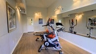 This single-floor fitness space is designed to support an effective and focused workout routine. The room features hardwood-style flooring that provides both durability and a clean, polished aesthetic. A large floor-to-ceiling mirror lines one wall, amplifying the sense of space and allowing users to monitor their form during exercise. The room is equipped with key cardio machines, including a spin bike and a treadmill, catering to a range of fitness preferences. Ambient string lighting and ceiling-mounted track lighting work together to create a well-lit yet welcoming atmosphere. Wainscoting along the lower walls adds a subtle architectural detail that elevates the room's overall appearance. Informational fitness guidelines are posted on the walls, contributing to the room's functional, safety-conscious design. A windowed door provides a connection to the exterior, allowing for natural light and ventilation. The space is thoughtfully laid out to maximize usability without feeling cluttered, making it a practical and inviting environment for daily exercise.