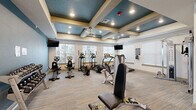 Located at 5803 SW Parc St in Bentonville, Arkansas, this fitness center offers a comprehensive workout facility spread across a single floor. The space is defined by its striking architectural ceiling design, featuring coffered panels finished in a bold teal-blue tone accented by cream-colored beams, recessed lighting, and ceiling fans that maintain air circulation throughout the room.

The facility is organized into distinct functional zones. The cardio area lines the windowed perimeter walls, housing multiple treadmills, elliptical trainers, and stationary bikes, with large windows providing ample natural light and outdoor views of the surrounding community. Wall-mounted flat-screen televisions are positioned throughout for entertainment during workouts.

The strength training section features a variety of selectorized machines, including cable systems, leg extension and curl equipment, and adjustable weight benches. A dedicated free weight area includes a full rack of dumbbells for versatile strength training. Neutral-toned carpet tiles cover the floors throughout, adding durability and comfort underfoot.

Entry to the fitness center is through elegant double glass-panel doors, flanked by built-in shelving for storing workout accessories. The overall ambiance is clean, modern, and well-lit, creating an inviting and functional environment for residents.