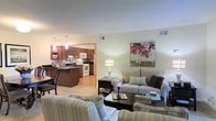This single-floor residence offers a thoughtfully designed open-concept layout that seamlessly connects the living and kitchen areas, creating a welcoming and functional living environment. The living room is comfortably furnished with neutral-toned sofas and accent chairs, centered around a dark wood coffee table. Warm ambient lighting from a classic table lamp and tasteful wall art add character and a homely feel to the space. The open flow into the kitchen makes the area feel expansive and well-connected. The kitchen is appointed with rich, warm-toned wood cabinetry that provides ample storage, complemented by dark granite countertops and a central island with seating. White appliances, including a range and built-in microwave, offer a clean contrast against the cabinetry. Pendant lighting hangs above the kitchen, enhancing both functionality and aesthetic appeal. Light-colored flooring runs throughout, tying the spaces together cohesively. The neutral color palette across the home, combined with soft lighting and an open layout, creates a bright, inviting ambiance that is both practical and comfortable for everyday living.