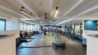 Located at 10050 E Harvard Ave in Denver, Colorado, this well-appointed fitness facility offers a generous, single-floor layout designed to accommodate a wide range of workout needs. The space is thoughtfully divided into distinct zones, each tailored for specific training disciplines.

The main workout floor features durable rubber flooring and hosts an extensive array of strength training equipment, including power racks, cable machines, weight benches, and free weights such as dumbbells and medicine balls. A dedicated functional training area, marked by turf flooring, provides space for sled drags, battle ropes, tire flips, and other dynamic exercises.

The cardio zone is lined with a full row of treadmills, stationary bikes, and elliptical machines positioned alongside large windows that invite ample natural light and outdoor views of the surrounding greenery. Full-length mirrors with teal accent walls and motivational signage enhance the energetic atmosphere throughout.

A separate studio area features wood-look flooring, floor-to-ceiling mirrors, and recessed lighting, making it ideal for group classes or individual training sessions. Distinctive curved brick partition walls add an architectural character to the space, while exposed beam ceilings and linear track lighting lend an industrial-modern aesthetic. An accessibility lift ensures the facility is fully accessible to all users.