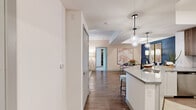 This thoughtfully designed single-floor residence offers a seamless blend of modern aesthetics and functional living. The open-concept kitchen is outfitted with stainless steel appliances, including a gas range, built-in microwave, and French door refrigerator, complemented by two-tone cabinetry — white lower cabinets paired with warm wood uppers — a tiled backsplash, and sleek quartz countertops that extend to a breakfast bar with pendant globe lighting overhead. A wide hallway connects the kitchen to the dining area, which features a tray ceiling, bold navy accent wall, and wood-look flooring that runs throughout the main living spaces. The living room is bright and airy, with a ceiling fan, large sliding glass door that invites natural light, and ample room for comfortable furnishings. The bedroom is a warm retreat with carpeted flooring, a generous window, and direct access to a spacious walk-in closet fitted with custom shelving, hanging rods, and a central ladder shelf unit. The bathroom is clean and contemporary, featuring wood-tone floating vanity cabinetry, an illuminated mirror, and a glass-enclosed shower. Completing the unit is a dedicated in-unit laundry area with a full-size washer and dryer and overhead shelving for storage.