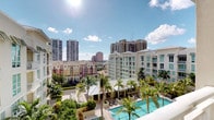 This single-floor residence is notably distinguished by its elevated vantage point, offering expansive views of a dynamic urban skyline punctuated by high-rise towers and mid-rise buildings. The outdoor perspective reveals a beautifully landscaped courtyard below, featuring a sparkling resort-style swimming pool surrounded by swaying palm trees and lounge seating, creating a tropical atmosphere in an urban setting. The surrounding neighborhood reflects a well-developed city environment with a mix of contemporary and classical architectural styles visible on the horizon. Under sunny skies with scattered clouds, the exterior setting exudes a bright, warm Florida ambiance. While interior photography is limited due to low lighting conditions, the residence is contained within a single level, suggesting a streamlined, efficient layout. The most compelling feature of this space is its connection to the outdoors—the elevated position provides commanding sightlines across the cityscape, making outdoor views a defining characteristic of the overall living experience. The combination of urban energy and resort-like amenities visible from the residence contributes to a distinctive and desirable atmosphere.