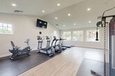 Located at 28 Fort Evans Rd NE in Leesburg, VA, this well-appointed fitness center offers a clean, modern environment designed to support a variety of workout routines. The single-story facility is organized into distinct zones, beginning with a bright entryway area finished in neutral tones with light wood-look flooring that flows throughout the space.

The main exercise area features vaulted ceilings with recessed lighting, creating an open and airy atmosphere. A wall-mounted flat-screen television is positioned at the front of the room for entertainment during workouts. Large windows along one wall invite abundant natural light and offer views of the surrounding outdoor greenery, enhancing the overall ambiance.

The cardio zone is well-equipped with multiple treadmills, elliptical trainers, and a stationary bike, all situated on protective rubber floor mats. A separate strength training area accommodates a multi-function cable machine, also placed on a rubber mat for safety and stability. A stainless steel waste receptacle is conveniently located nearby.

The entryway includes two access doors and HVAC units, ensuring comfortable climate control year-round. Security features such as a mounted camera and exit signage are also present. The facility's bright white walls, crisp white trim, and minimalist design create a professional and inviting fitness environment.