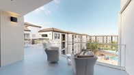 This single-level rooftop terrace is a well-appointed outdoor amenity space set atop a Mediterranean-inspired residential building. The terrace is finished with a smooth, light blue-grey coated floor that runs throughout, giving the entire level a clean, cohesive aesthetic that complements the white stucco exterior walls and terracotta tile rooflines visible throughout the surrounding community.

The space is organized into several distinct zones. An open-air lounge area features contemporary upholstered armchairs and low side tables arranged to take full advantage of the panoramic views, with glass-panel railings ensuring unobstructed sightlines across the palm-tree-lined grounds below. A covered outdoor lounge section, fitted with a ceiling fan, provides shade and shelter while maintaining the open-air feel through large floor-to-ceiling sliding glass doors that connect seamlessly to the interior.

An outdoor bar and kitchen area serves as a social hub, anchored by a substantial stone-top bar counter with woven high-back barstools. A built-in grill station and a dark-tiled countertop with a sink complete the outdoor culinary setup, alongside an outdoor television mounted nearby for entertaining. A sun lounger positioned near the glass doors invites relaxation while capturing natural light throughout the day. The overall ambiance is resort-inspired, bright, and open.
