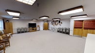 This single-floor space functions as a versatile multipurpose room well-suited for gatherings, presentations, or group activities. The open, carpeted floor area provides ample room for flexible arrangements, making it adaptable for a variety of uses. Overhead illumination is provided by a combination of skylight panels and several large rectangular flush-mount light fixtures, ensuring the room is consistently and evenly lit throughout. The neutral-toned walls and light carpet contribute to a clean, functional aesthetic. Along one wall, a flat-screen television is positioned atop a wooden media console, accompanied by a tall wooden armoire-style cabinet for storage. A collection of woven baskets and decorative circular wall hangings adds a warm, casual character to the space. Several folding chairs are neatly lined along the perimeter, indicating the room's capacity to accommodate seated groups. A doorway is visible along the right wall, providing access to adjacent areas. Overall, the room presents a practical and organized environment designed to support community or organizational activities with ease.