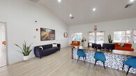 This thoughtfully designed single-floor community clubhouse blends modern aesthetics with functional amenities across several distinct areas. The main entrance opens into a bright, airy reception and lobby area distinguished by soaring vaulted ceilings and light hardwood-style flooring. Large windows and glass double doors invite abundant natural light, offering views of the surrounding exterior. A structured front desk workstation with a bold geometric-patterned front panel serves as the central hub, complemented by teal velvet accent chairs and a sleek navy channeled sofa. Curated artwork featuring dramatic landscape photography adds personality to the crisp white walls.

Adjacent to the reception is a welcoming lounge seating area with woven accent chairs and a glass coffee table, ideal for informal gatherings. The space flows into a vibrant recreation room equipped with a foosball table, eclectic seating arrangements, and a kitchenette featuring open shelving, a sink, and counter space — all set against a striking decorative wall panel. A dedicated fitness center rounds out the amenities, outfitted with treadmills, a stationary bike, a cable weight machine, and a wall-mounted television, set on durable carpet flooring. Throughout, the cohesive mid-century modern design palette creates an inviting and energetic atmosphere.
