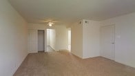 This single-floor space presents a clean, functional layout with neutral cream-colored walls and wall-to-wall beige carpeting throughout, creating a cohesive and understated aesthetic. The main room is generously proportioned, providing ample open floor space suitable for a variety of furniture arrangements. A ceiling fan with an integrated light fixture is centrally mounted, offering both air circulation and ambient lighting.

A built-in closet with bifold doors is visible along one wall, providing convenient storage directly within the living area. An HVAC vent is positioned on the upper wall, ensuring climate control throughout the space. The room opens into a connecting hallway or adjoining area, visible through a wide doorway, suggesting a practical flow between rooms. A second doorway on the right side of the space leads to an additional room, further highlighting the functional connectivity of the layout.

The overall ambiance is simple and move-in ready, with a neutral palette that offers flexibility for personalization. The absence of ornate detailing emphasizes practicality, making this space well-suited for straightforward, everyday living.