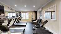 This fitness center offers a thoughtfully organized workout environment designed to accommodate a range of exercise needs. The space features a row of treadmills along one side, providing ample cardio options for residents or members. On the opposite side, a multi-tiered dumbbell rack holds a comprehensive set of free weights alongside an adjustable workout bench, supporting strength training routines of varying intensity. The flooring consists of durable rubber matting, providing both safety and comfort during workouts. Natural light filters in through multiple windows, lending the room an open and energizing atmosphere, while recessed ceiling lights ensure consistent illumination throughout. Crown molding along the ceiling adds a refined architectural touch, elevating the overall aesthetic beyond a typical gym setting. A wall-mounted air conditioning unit supports climate control, keeping the environment comfortable year-round. Motivational athletic posters and wall graphics contribute to an active and focused ambiance. The layout is efficient and well-organized, allowing users to move freely between equipment without congestion, making the space functional for both individual and simultaneous use.