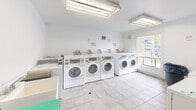 Located at 1200 Courtland Ave E in Kitchener, Ontario, this shared laundry facility offers a practical and functional space designed to serve building residents efficiently. The room features white walls and light tile flooring throughout, contributing to a clean and bright atmosphere. Overhead fluorescent lighting ensures the space is well-lit at all times, supplemented by natural light from a large window that looks out onto the surrounding grounds and greenery.

The facility is equipped with multiple front-loading washers and top-loading dryers arranged in rows along the walls, providing ample capacity for shared use. A utility sink is also present, offering added convenience for hand-washing or pre-treatment of garments. A waste bin and a small utility table provide additional functional support within the space.

The room connects to an adjacent corridor through a doorway, allowing for straightforward access from the building's common areas. Exposed plumbing and ventilation elements along the upper walls reflect the utilitarian nature of the space. Notices and signage are posted on the walls, providing usage guidelines for residents. Overall, the laundry room is a well-equipped, no-frills communal facility designed to meet the everyday laundry needs of building occupants.