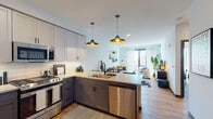 Located at 131 Mamaroneck Ave in White Plains, NY, this contemporary residence offers a thoughtfully designed open-concept layout finished with warm hardwood flooring throughout. The kitchen is a standout feature, equipped with stainless steel appliances including a gas range, microwave, and dishwasher, complemented by two-tone cabinetry — crisp white uppers paired with charcoal lower cabinets — and light quartz countertops. A peninsula with seating provides a natural gathering point, while statement pendant lights with gold interiors add a touch of industrial-modern flair. The kitchen flows seamlessly into the living area, which is bathed in natural light from floor-to-ceiling windows offering expansive views of the White Plains skyline. The bedroom features a bold slate-blue accent wall, large windows with city views, and direct access to a well-organized walk-in closet. The bathroom is finished with clean white subway tile in the tub surround, stone-look tile flooring, a shaker-style vanity, and matte black fixtures throughout, conveying a refined, contemporary aesthetic. The overall ambiance is modern and urban, with a cohesive design palette that balances functionality and style.