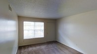 This single-floor residential space presents a straightforward and functional layout suited for comfortable everyday living. The interior is finished with wall-to-wall carpet flooring in a neutral tone, contributing to a quiet and cozy atmosphere throughout. The walls are painted in a soft, light neutral shade, keeping the space feeling open and easy to furnish. A textured popcorn ceiling adds a classic touch to the overhead finish, consistent with the overall understated aesthetic of the unit. Natural light enters through a standard window fitted with horizontal blinds, allowing for adjustable light control and a view to the exterior. A doorway with a paneled door provides access to an adjoining space, supporting a practical flow between areas. The room is currently unoccupied, making it easy to assess its proportions and envision a variety of furniture arrangements. The clean, minimal design of the space provides a neutral canvas for personalization. Overall, the space offers a functional and accessible residential environment with simple, durable finishes throughout.