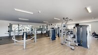 This expansive fitness center offers a generously sized, single-level layout designed to accommodate a comprehensive range of workout needs. The space is defined by crisp white walls, smooth ceilings with rows of bright linear fluorescent lighting, and light-toned wood-look flooring that flows throughout the majority of the facility. A dedicated strength training zone features a wide array of selectorized cable machines, multi-station cable crossovers, free weight racks with dumbbells, weight benches, and functional training rigs, all positioned atop durable rubberized flooring for added safety and performance. Full-length mirrors line the strength area walls, enhancing the sense of openness while allowing users to monitor their form. The cardio section is equipped with treadmills, elliptical trainers, rowing machines, and upright bikes, positioned near large windows that allow natural light to fill the space and offer views of the surrounding outdoors. A separate lounge and stretching area provides a relaxed atmosphere with comfortable seating, a dining table with chairs, a wall-mounted television, and direct access to an exterior door. The overall ambiance is clean, modern, and functional, creating an inviting environment suited for both casual exercisers and dedicated fitness enthusiasts.