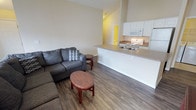 This single-floor unit offers an open-concept layout that seamlessly connects the living area and kitchen, creating a spacious and functional environment. The living area features warm hardwood-style flooring that extends throughout the space, complemented by neutral-toned walls that lend a clean, versatile aesthetic. Notably high vaulted ceilings add a sense of openness and airiness to the room. The living space comfortably accommodates a sectional-style sofa and accent tables, providing a welcoming area for relaxation.

The kitchen is positioned adjacent to the living area and is defined by a peninsula counter with a light-toned laminate surface, offering additional workspace and serving as a natural divider between the two zones. White cabinetry lines the kitchen walls, providing ample storage, while standard appliances — including a range, microwave, and refrigerator — are integrated into the layout. A double-basin sink sits beneath upper cabinets, completing a practical and efficient cooking space.

A hallway visible from the main living area leads to additional rooms, suggesting a thoughtfully organized floor plan. Overall, the space maintains a bright, neutral ambiance suited for everyday living.