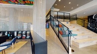 This expansive single-floor space combines a dynamic mix of design elements to create a visually engaging, multi-functional environment. The entrance corridor features textured dark walls with a moss-like finish, natural hardwood flooring, and a narrow glazed door that frames street views beyond. A polished concrete floor runs through the main lobby, where Foxtail Coffee Co. anchors one end beneath bold graphic wall murals rendered in black and white. A wide, light wood staircase with sleek black metal railings leads to a mezzanine-level lounge area, creating vertical interest throughout the space. Cascading metallic chain curtains serve as dramatic yet functional dividers between zones. The central lounge area features vivid red accent walls, a large sectional sofa, mounted television, and hardwood flooring. Adjacent to this is a dining and collaboration zone beneath a striking, vibrantly painted ceiling mural bursting with color. Glass-enclosed meeting pods line one corridor, while a generous window wall dressed in sheer white curtains floods a bar-height communal work counter with natural light and exterior views. The overall ambiance is energetic, creative, and contemporary.