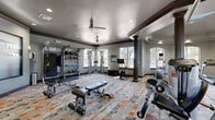 This fitness center offers a thoughtfully designed workout space spread across a single open floor, providing ample room for a comprehensive range of exercise equipment and activities. The space is anchored by distinctive dark decorative columns and finished with rich crown molding, lending a refined architectural character that sets it apart from a typical gym environment. The ceiling features recessed lighting and dual ceiling fans, ensuring the space remains well-lit and comfortable throughout.

The colorful, patterned carpet tiles in shades of gray, orange, and white add a dynamic visual element to the floor while providing cushioning underfoot. Natural light streams in generously through multiple arched and rectangular windows, offering views of surrounding greenery and creating a bright, welcoming atmosphere.

Equipment is varied and plentiful, including a Hoist multi-station cable machine, a TRX suspension training system, a freestanding rack with medicine balls and kettlebells, adjustable weight benches, a dumbbell rack, and a plyometric box. Cardio options include a Precor elliptical, treadmills, a stationary bike, and a cycling bike. Full-length mirrors along one wall enhance the sense of space and allow users to monitor their form. The overall ambiance is clean, organized, and motivating, making this fitness center a highly functional and attractive amenity.