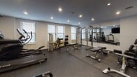 Located at 100 Jersey St in Boston's Fenway neighborhood, this well-equipped fitness center offers a comprehensive range of workout options within a single, thoughtfully organized space. The room features durable rubber flooring throughout, providing a safe and functional surface for all types of exercise.

Cardio equipment includes True-brand treadmills and an elliptical machine, each equipped with individual digital displays. A wall-mounted flat-screen television adds an entertainment option for those using the cardio stations. Natural light enters through multiple windows fitted with blinds, complementing the recessed ceiling lighting that keeps the space bright and well-illuminated.

The strength training area is anchored by a full power rack with an adjustable bench, suitable for a variety of barbell exercises. A dedicated free weight section includes a tiered dumbbell rack stocked with multiple weight increments, alongside additional benches and a cable/functional trainer machine. A large wall mirror spans a significant portion of one wall, enhancing the sense of space and allowing for form checks during exercises.

A stationary bike and rowing machine round out the cardio offerings, while a water cooler and AED device are conveniently accessible. A separate door leads to an adjacent area, likely housing restroom or changing facilities. The overall ambiance is clean, modern, and purpose-built for fitness.