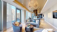 Located in the heart of Center City Philadelphia on Sansom Street, this single-floor living space is a masterclass in upscale interior design. The open-plan layout is anchored by a dramatic freestanding cylindrical fireplace encased in polished bronze and glass, serving as a striking focal point that commands attention from every angle.

The living area features elegant herringbone hardwood flooring, a luminous gold-leaf tray ceiling, and a sculptural branch-inspired chandelier that casts a warm, ambient glow. Walls of floor-to-ceiling windows flood the interior with abundant natural light while framing sweeping panoramic views of the Philadelphia skyline. A private outdoor terrace extends the living space, accessible through large glass doors.

A bold, atmospheric blue watercolor mural adds an artistic dimension to one wall, complemented by refined furnishings in soft grays, blues, and warm gold tones. The integrated bar area features a warm wood slat ceiling, a sleek marble countertop, brass pendant lighting, and velvet upholstered barstools—all set against the backdrop of the city skyline.

Vertical LED wall sconces, panel molding, and layered lighting throughout create a cohesive, polished ambiance. Every detail of this space speaks to sophisticated, contemporary urban living at its finest.