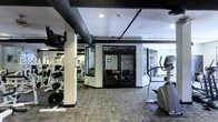 This fitness center offers a comprehensive workout environment thoughtfully organized across a single level. The space is defined by its industrial aesthetic, with exposed ductwork running along a dark ceiling that contrasts sharply with the lighter walls and support columns, creating a dynamic and energetic atmosphere.

The facility is divided into distinct workout zones, separated by a set of sleek, black-framed glass double doors that allow visibility throughout the space while providing functional separation. On one side, a well-stocked strength training area features weight racks, barbells, and a variety of resistance machines arranged to maximize floor space and ease of movement. The opposite section is dedicated to cardiovascular equipment, with stair climbers and additional machines positioned near large windows that invite natural light into the space.

Underfoot, durable patterned carpet tiles cover the floor throughout, providing both comfort and practicality for a high-traffic fitness environment. Structural columns are integrated throughout the layout, reinforcing the industrial character of the space. The overall design balances utility and aesthetics, offering a well-lit and organized facility suitable for a wide range of fitness routines, from strength-focused workouts to cardio training sessions.