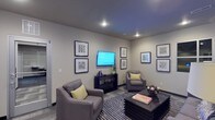This single-floor space presents a thoughtfully designed lounge or sitting area with a clean, modern aesthetic. The room is anchored by two plush gray armchairs accented with chartreuse throw pillows, arranged around a dark, slatted rectangular coffee table that serves as a stylish focal point. A wall-mounted flat-screen television is positioned above a matching dark console unit, creating a natural media wall. The neutral gray walls provide a calm backdrop for a carefully curated gallery of framed artwork, arranged symmetrically on two walls to add visual interest without overwhelming the space. Recessed ceiling lighting illuminates the room evenly, complementing the cool-toned gray ceiling. A striped area rug in charcoal and silver tones defines the seating zone and adds texture underfoot. Decorative objects, including a bowl of spherical accents and a small plant, contribute to the polished yet approachable atmosphere. The space conveys a sense of comfort and functionality, making it well-suited as a communal lounge, waiting area, or social gathering space.