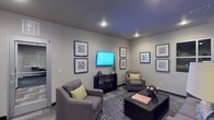 This single-floor space presents a thoughtfully designed lounge or sitting area with a clean, modern aesthetic. The room is anchored by two plush gray armchairs accented with chartreuse throw pillows, arranged around a dark, slatted rectangular coffee table that serves as a stylish focal point. A wall-mounted flat-screen television is positioned above a matching dark console unit, creating a natural media wall. The neutral gray walls provide a calm backdrop for a carefully curated gallery of framed artwork, arranged symmetrically on two walls to add visual interest without overwhelming the space. Recessed ceiling lighting illuminates the room evenly, complementing the cool-toned gray ceiling. A striped area rug in charcoal and silver tones defines the seating zone and adds texture underfoot. Decorative objects, including a bowl of spherical accents and a small plant, contribute to the polished yet approachable atmosphere. The space conveys a sense of comfort and functionality, making it well-suited as a communal lounge, waiting area, or social gathering space.