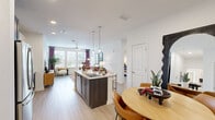 This thoughtfully designed single-floor residence showcases a bright, open-concept layout that seamlessly connects the kitchen, dining, and living areas. The kitchen serves as the centerpiece, anchored by a large island with granite countertops, a undermount sink, and pendant lighting overhead. Dark espresso cabinetry contrasts elegantly with white upper cabinets, while stainless steel appliances—including a French door refrigerator and microwave—add a polished, modern touch. A subway tile backsplash completes the look.

The dining area accommodates a round wood table with amber-toned chairs, positioned near the entry. The living room benefits from generous natural light through large windows dressed with bold plum curtains and a glass-paned exterior door, creating an airy and welcoming atmosphere. Light wood-tone flooring runs consistently throughout, tying all areas together cohesively.

The bedroom is a serene retreat with ample window light and direct access to the en-suite bathroom, which features a glass-enclosed walk-in shower, dual-sink vanity with granite countertops, and a backlit mirror. A spacious walk-in closet with wire shelving provides generous storage. A dedicated in-unit laundry area with a full-size washer and dryer adds everyday convenience. The overall ambiance is clean, contemporary, and functional.