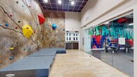 Located at 2121 S Hiawassee Rd in Orlando, Florida, this dynamic fitness and recreation facility combines athletic functionality with bold artistic expression across a single, well-appointed floor. The space is immediately distinguished by its striking interior design, where large-scale graffiti-style murals in vivid blues, reds, greens, and yellows energize the walls throughout.

The standout feature is a towering bouldering wall adorned with colorful climbing holds in red, yellow, blue, and green. The wall rises to impressive heights beneath soaring ceilings, with protective crash mats laid out below to ensure safe landings. Light-toned stone tile flooring runs through this area, complemented by a dark, grid-patterned ceiling with recessed lighting that adds a modern contrast.

Adjacent to the climbing area, the fitness floor is expansive and fully equipped with a comprehensive selection of SportsArt cardio machines, including treadmills, stationary bikes, and step trainers, alongside a wide array of strength-training equipment, cable machines, free weights, and benches. Rubber flooring throughout the gym provides durability and safety, while wall-mounted flat-screen TVs offer entertainment during workouts.

The facility's entrance corridor features an elegant, contrasting aesthetic with ornate patterned tile flooring, crisp white walls, decorative molding, and classic chandelier lighting — creating a refined welcome before transitioning into the energetic fitness environment beyond.