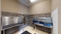 This single-level pet wash facility is purpose-built for grooming and bathing pets with efficiency and cleanliness in mind. The room is finished with large-format, light-toned porcelain floor tiles and a bold horizontal band of textured stone-look wall tiles that run along the lower half of all walls, adding a polished, durable aesthetic suited to a high-use environment.

At the core of the space are two stainless steel grooming surfaces: a perforated-top grooming table ideal for drying and brushing, and a dedicated wash basin equipped with a coiled blue hose, a mounted handheld sprayer, and a central drain for efficient water management. A wall-mounted blow dryer attachment and shelving brackets offer additional grooming utility. Anti-fatigue rubber mats are positioned at key standing areas, enhancing comfort during use.

Overhead, recessed fluorescent lighting provides bright, even illumination throughout the room. A glass-paneled entry door allows natural light to filter in, and posted usage guidelines are clearly visible near the entrance. The neutral taupe wall color, combined with the stone-tile detailing, creates a clean and functional environment that balances practicality with a well-designed finish.