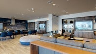 This thoughtfully designed single-floor amenity space offers a sophisticated, open-concept layout that seamlessly blends social and recreational areas. Light hardwood flooring runs throughout, creating a warm and cohesive foundation that ties the various zones together.

At the heart of the space stands a striking white brick fireplace column with a sleek, modern firebox, flanked by a built-in dark wood entertainment wall complete with open shelving, curated décor, and integrated media displays. Plush sectional seating and accent chairs invite relaxed lounging, while a secondary seating area with dark upholstered sofas and a wood coffee table offers an additional gathering spot.

The full demonstration kitchen is a standout feature, boasting dark cabinetry, a bold textured tile backsplash against a deep navy accent wall, a large central island with bar seating, and professional-grade appliances beneath an industrial range hood.

A dining area with rich wood-paneled walls and blue upholstered chairs provides an elegant setting for shared meals, while intimate semi-private dining nooks with textured white accent walls offer a more secluded atmosphere. Floor-to-ceiling windows flood the space with natural light and frame views of an outdoor pool and lounge deck. A built-in window bench with navy cushions serves as a quiet retreat. The floorplan also reveals a dedicated game area with a billiards table, rounding out this versatile and well-appointed communal space.