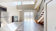 Located at 5250 Liberty Avenue in Pittsburgh's Bloomfield neighborhood, this two-story loft-style residence is thoughtfully designed across two levels. The main floor centers on an open-concept kitchen and living area with soaring double-height ceilings that flood the space with natural light through large clerestory windows. Exposed spiral ductwork lends an industrial character, while light-toned hardwood flooring runs throughout. A sleek, freestanding fireplace anchors the living area, and floor-to-ceiling sliding glass doors open directly onto a private balcony with open urban views. The well-appointed kitchen features rich, dark cabinetry, white quartz countertops, stainless steel appliances, a built-in wine rack, and a movable stainless steel island. A striking open-riser staircase with wooden treads and a black metal mesh railing leads to the upper loft level. Built-in closets provide ample storage on the main floor, alongside a dedicated laundry area and a den. The full bathroom includes a tub-shower combination with subway tile surround and warm hardwood flooring. Upstairs, the loft offers a flexible additional space with carpeted flooring and a walk-in closet with built-in wire shelving. The overall aesthetic is modern and urban with warm, neutral finishes throughout.
