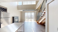 Located at 5250 Liberty Avenue in Pittsburgh's Bloomfield neighborhood, this two-story loft-style residence is thoughtfully designed across two levels. The main floor centers on an open-concept kitchen and living area with soaring double-height ceilings that flood the space with natural light through large clerestory windows. Exposed spiral ductwork lends an industrial character, while light-toned hardwood flooring runs throughout. A sleek, freestanding fireplace anchors the living area, and floor-to-ceiling sliding glass doors open directly onto a private balcony with open urban views. The well-appointed kitchen features rich, dark cabinetry, white quartz countertops, stainless steel appliances, a built-in wine rack, and a movable stainless steel island. A striking open-riser staircase with wooden treads and a black metal mesh railing leads to the upper loft level. Built-in closets provide ample storage on the main floor, alongside a dedicated laundry area and a den. The full bathroom includes a tub-shower combination with subway tile surround and warm hardwood flooring. Upstairs, the loft offers a flexible additional space with carpeted flooring and a walk-in closet with built-in wire shelving. The overall aesthetic is modern and urban with warm, neutral finishes throughout.