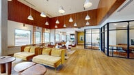 Located at 500 West Camelback Road in Phoenix, AZ, this thoughtfully designed single-floor community space blends contemporary aesthetics with warm, inviting materials. The layout spans a generous footprint and is unified by wide-plank light hardwood flooring and dramatic, soaring ceilings clad in rich wood slatted paneling that add texture and warmth throughout.

The main lounge area is anchored by plush mustard-yellow velvet sofas and tan leather armchairs arranged around sculptural wood and dark accent tables, creating an inviting setting for relaxation or casual gathering. Clusters of white cone-shaped pendant lights hang gracefully from the elevated ceiling, complementing the natural light that pours in through large picture windows along the perimeter. A wall-mounted flat-screen display is prominently featured in the lounge zone.

A dedicated dining and collaboration area features a long communal table with seating for multiple guests, set against a striking dark tile backsplash and built-in shelving. Decorative lattice wood screens provide visual separation while maintaining an open, airy feel. An enclosed glass-walled room with navy steel-framed doors offers a private meeting or conference space. A built-in bench seat with cushions runs below a row of windows, providing an additional casual seating nook. The space also includes a bathroom, completing this well-appointed communal environment.