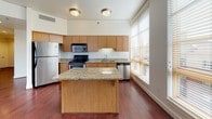 This single-floor residence offers a thoughtfully designed layout that balances function and comfort throughout its well-appointed spaces. The living area is a standout, featuring rich, deep-toned hardwood flooring that extends through the main living spaces. Large windows fitted with wood-toned blinds flood the room with natural light and provide views of the surrounding neighborhood, while a sliding glass door opens to a private balcony. The kitchen is well-equipped for daily living, showcasing warm maple cabinetry, granite countertops, and a full suite of stainless steel appliances including a range, over-the-range microwave, refrigerator, and dishwasher. A central granite-topped island provides additional prep space and casual seating. The bedrooms are carpeted for comfort, with clean white walls and ample ceiling ventilation. Two bathrooms serve the residence: one features a granite vanity, white cabinetry, and a tub-shower combination, while the second offers a glass-enclosed walk-in shower and toilet in a bright, neutrally finished space. Throughout, recessed lighting and flush-mount ceiling fixtures provide consistent illumination. The overall palette of warm wood tones, crisp white walls, and granite accents creates a cohesive and inviting atmosphere.
